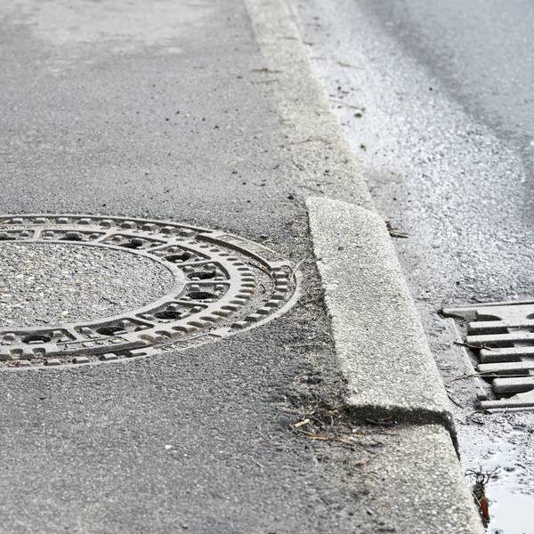 Ein Gehweg mit einem Gullideckel. Ein Stein der Bordsteinkante hat sich gesenkt.