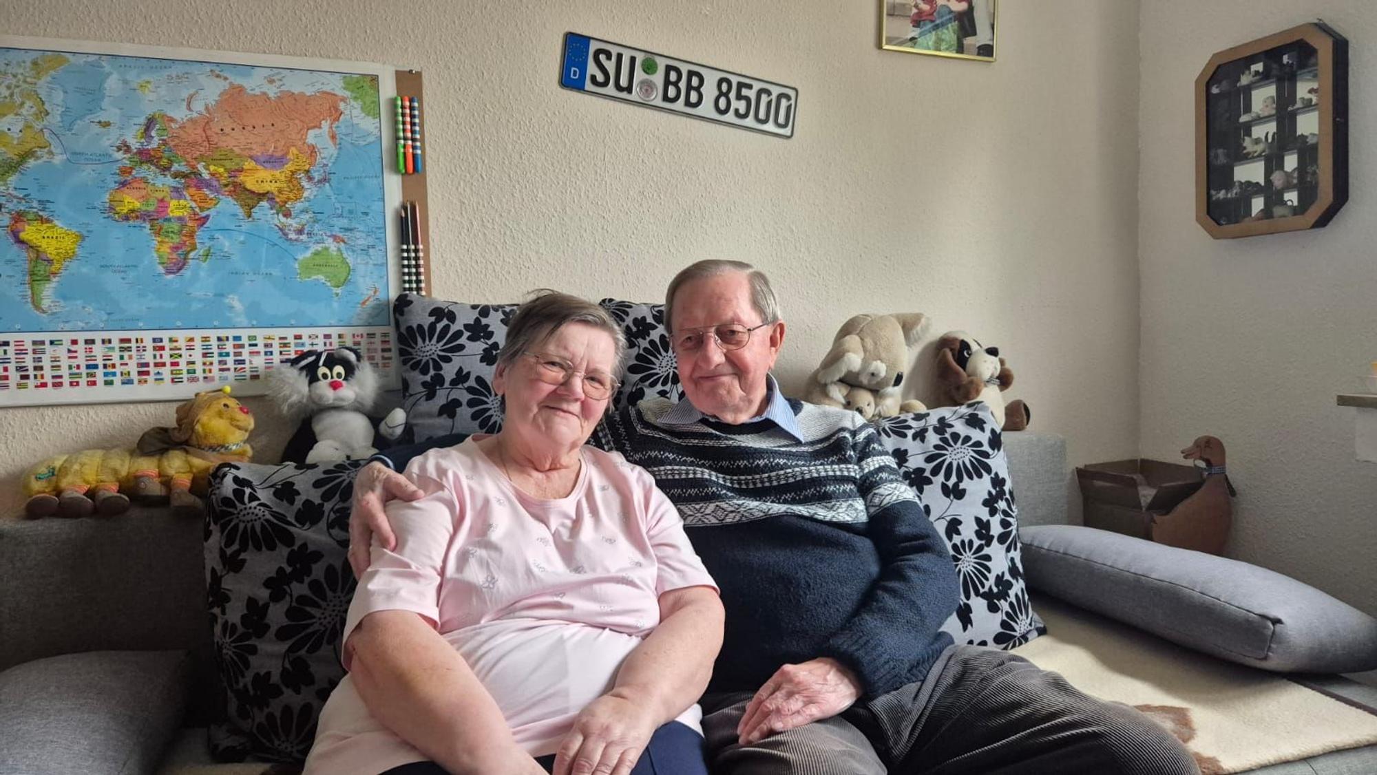 Ein älteres Ehepaar sitzt auf einem Sofa, auf dessen Lehne Kuscheltiere sitzen.