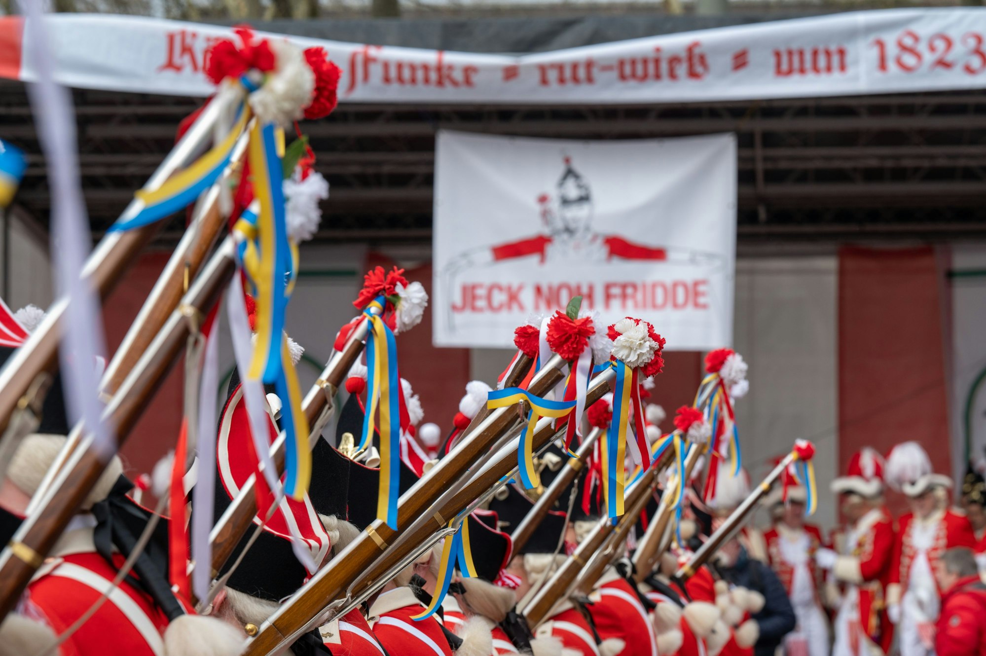26.02.2022, Köln: gelb- blaue Bändchen in den Nationalfarben der Ukranie an den Knabbüß genannten Holzgewehren der Roten Funken. Transparant "Jeck noh Fridde" gegen den Krieg Russlands in der Ukraine. Funkenbiwak der Roten Funken am Neumarkt. Foto: Uwe Weiser