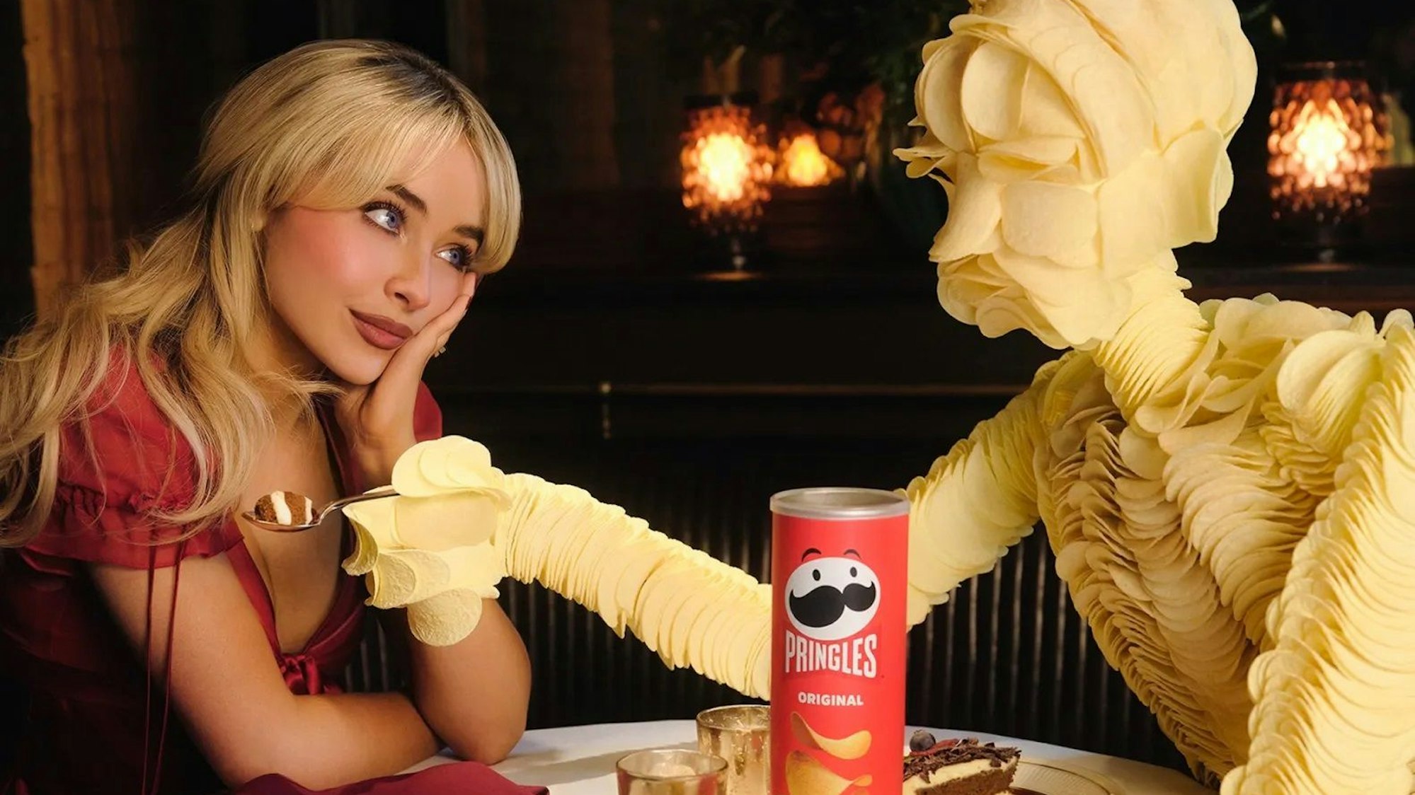 Sabrina Carpenter sitzt mit aufgebeugten Armen an einem Restauranttisch. Ihr gegenüber ein Mann aus Pringle-Chips.