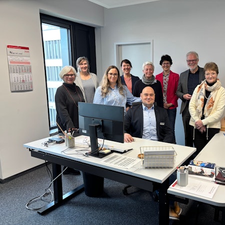 Eine Gruppe Menschen steht hinter Schreibtischen in einem Büroraum. Ein Mann sitzt vor ihnen und hat seine Hand auf einem der Schreibtische abgestützt.