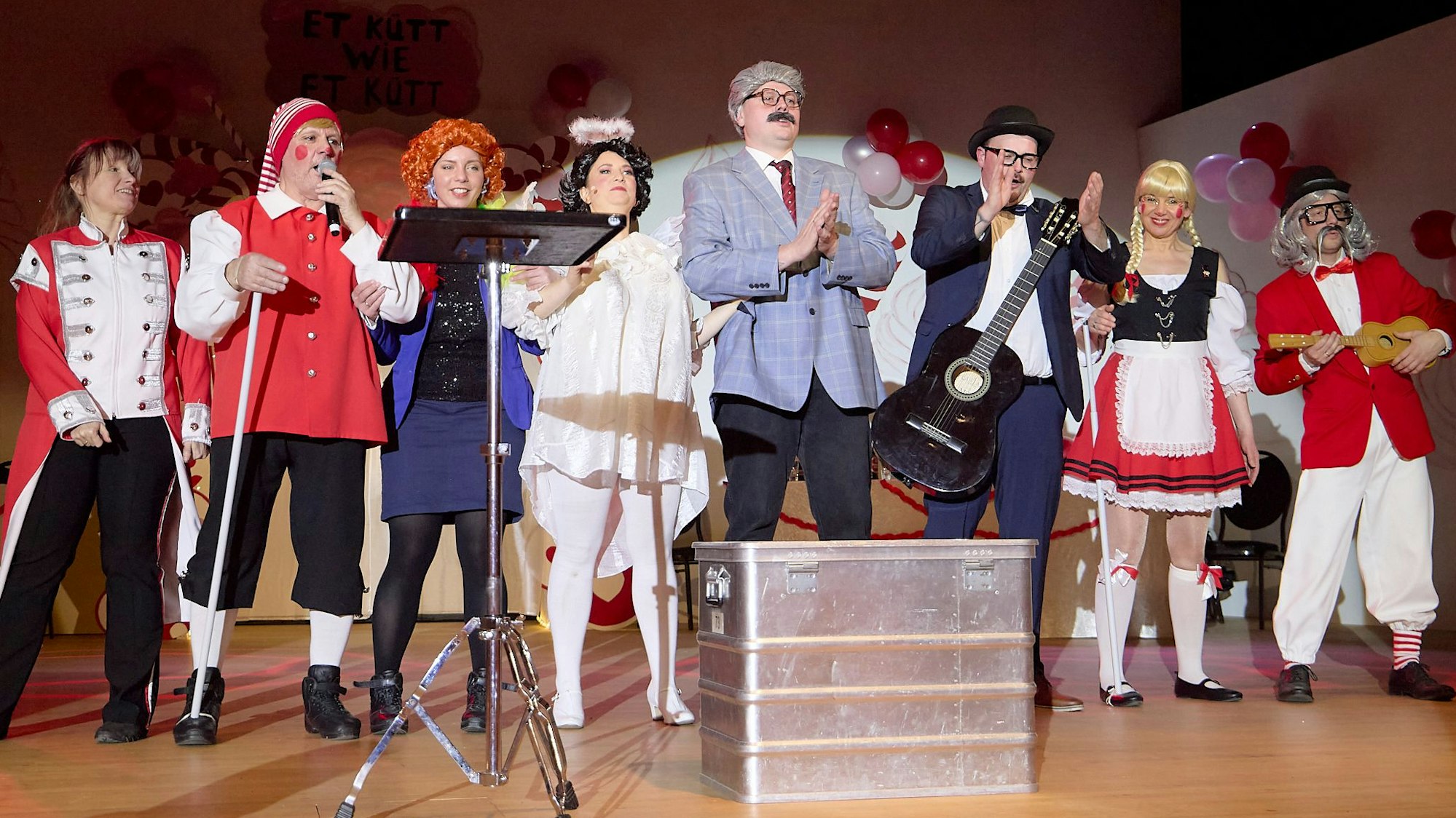 Acht Karnevalistinnen und Karnevalisten haben sich als Größen aus dem Kölner Karneval verkleidet und stehen auf einer Bühne.