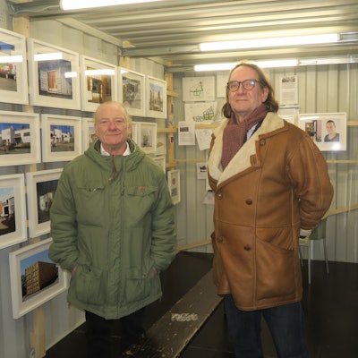 Zwei Männer stehen in einem Container, in dem Fotos an den Wänden hängen.