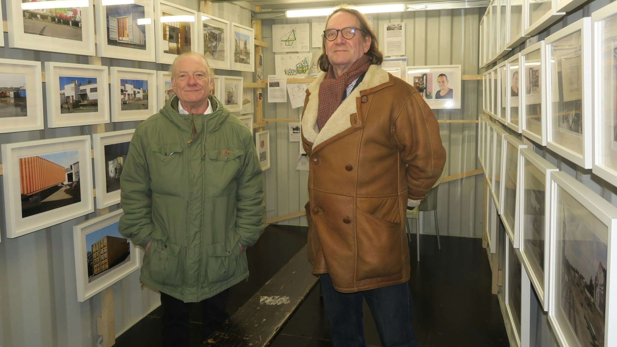 Zwei Männer stehen in einem Container, in dem Fotos an den Wänden hängen.