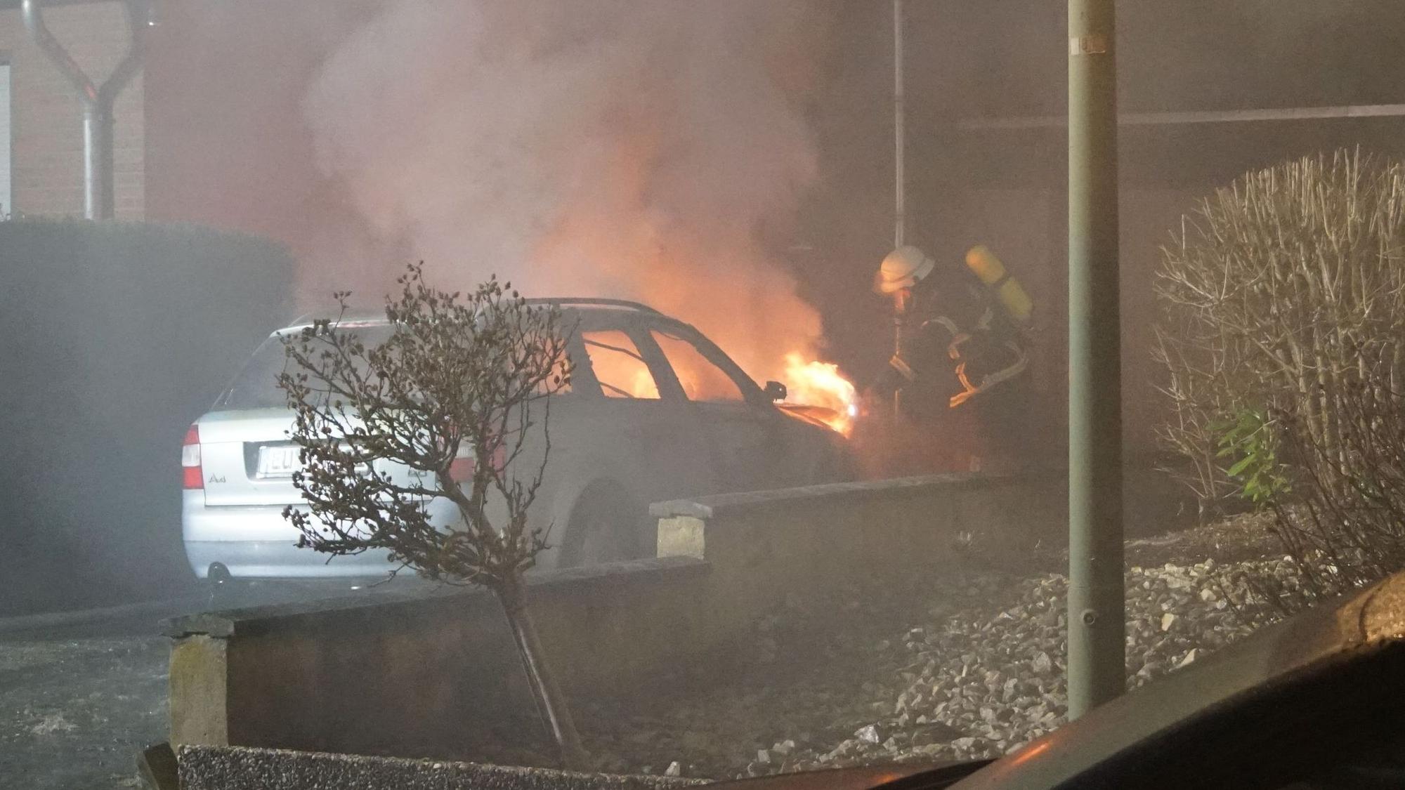 Ein Auto steht in der Einfahrt zu einem Haus. Aus der Motorhaube schlagen Flammen, eine Einsatzkraft der Feuerwehr beginnt mit den Löscharbeiten.