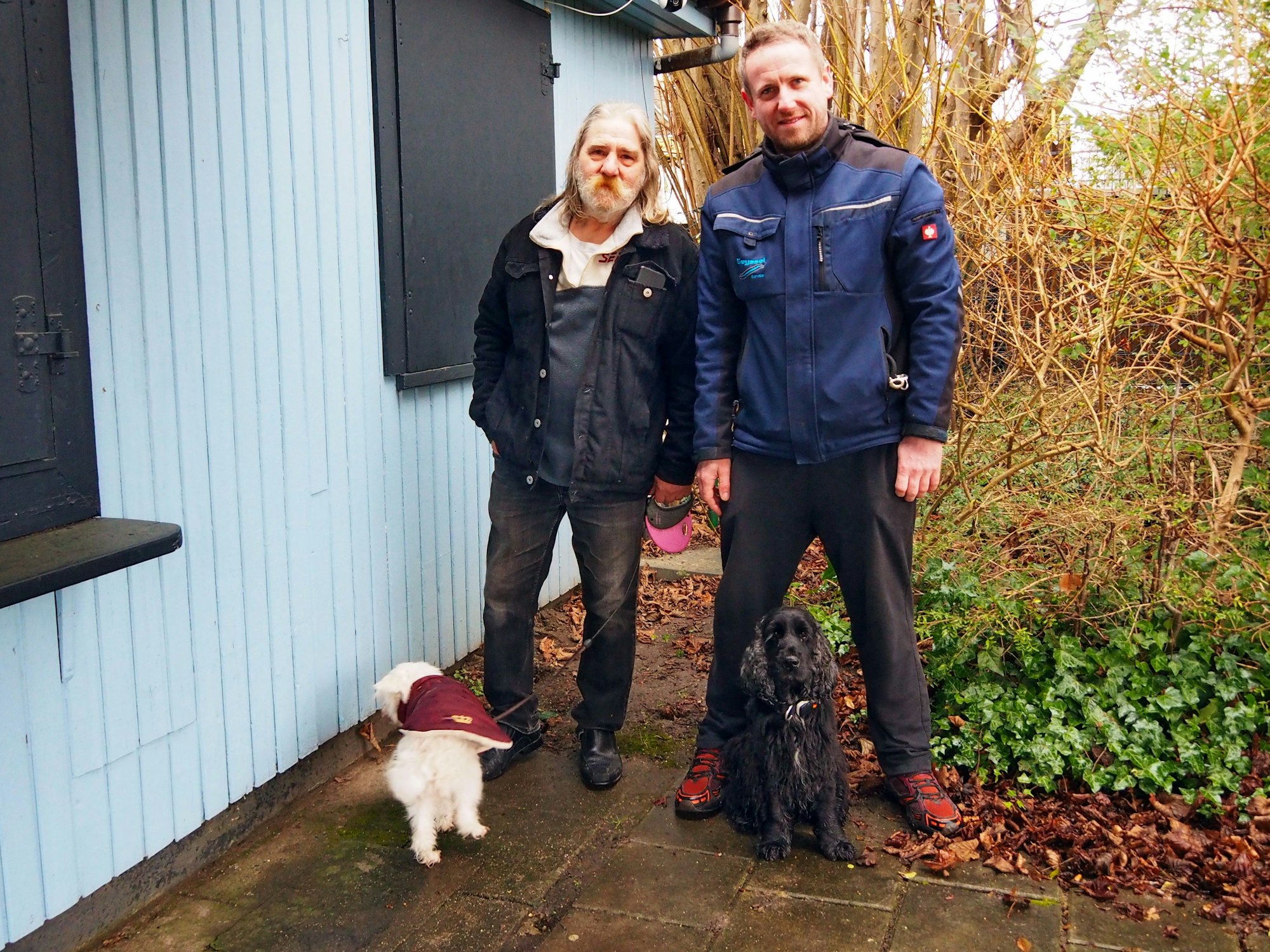 Zwei Männer stehen mit ihren beiden Hunden neben der Laube einer Kleingartenanlage.