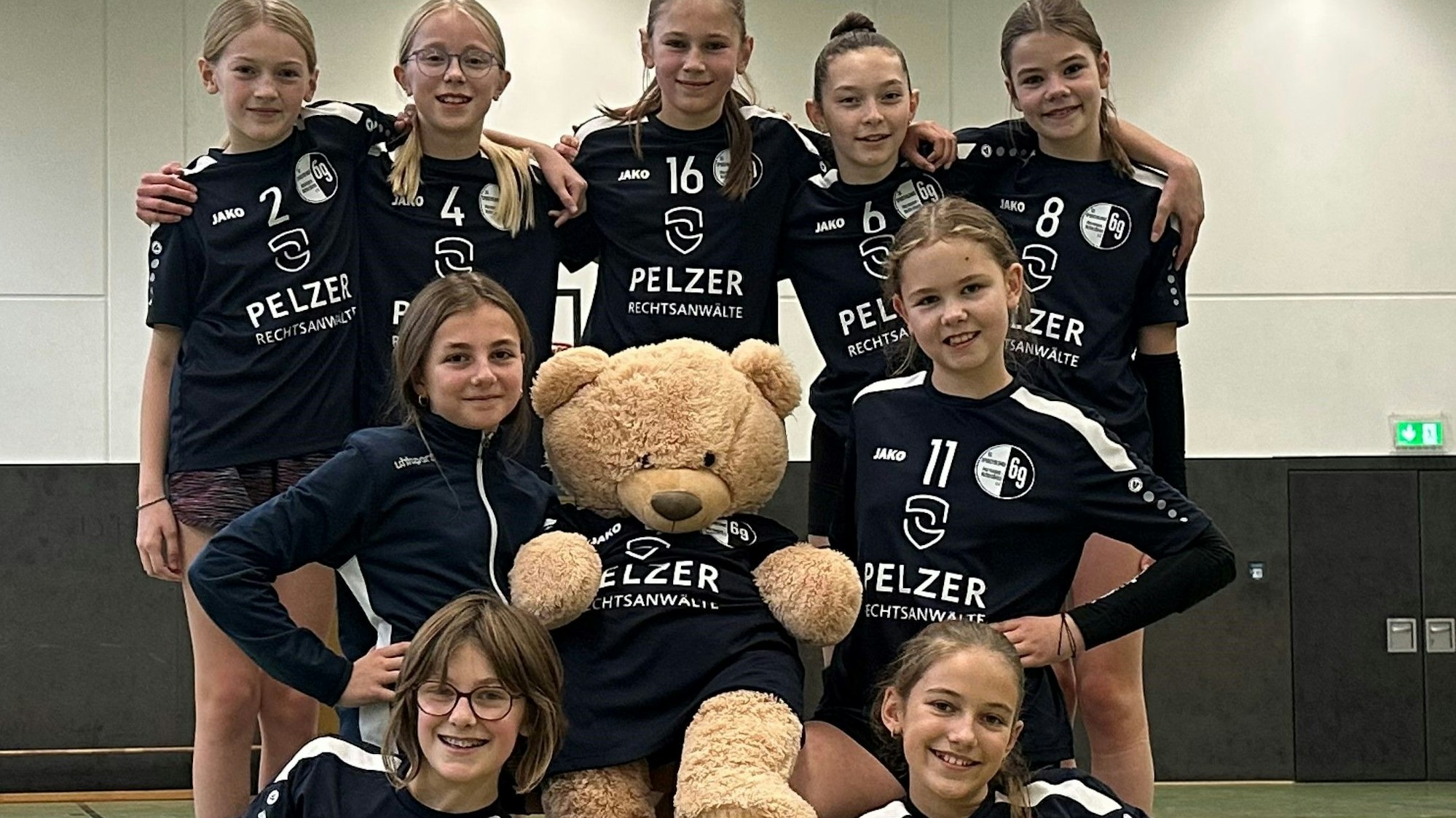 Mädchen in Vollyballanzügen haben sich um einen großen Teddybären aufgestellt.