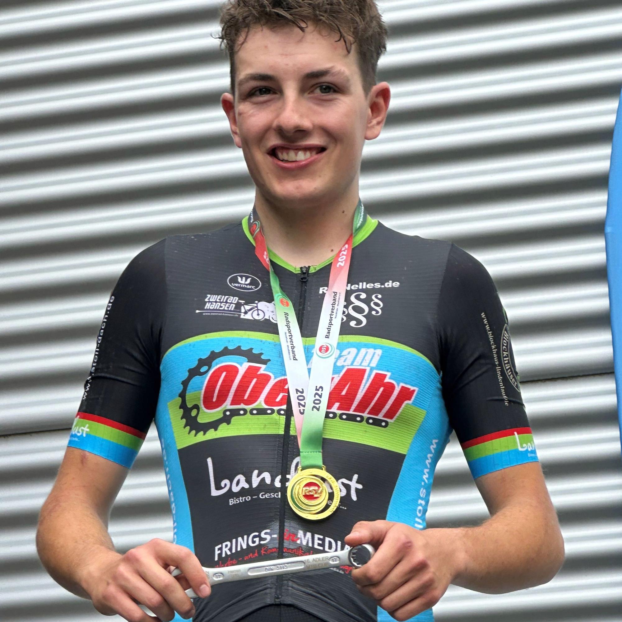 Phil Sawinsky steht in einem Fahrradtrikot und mit Medaille um den Hals vor einer Wellblechwand.