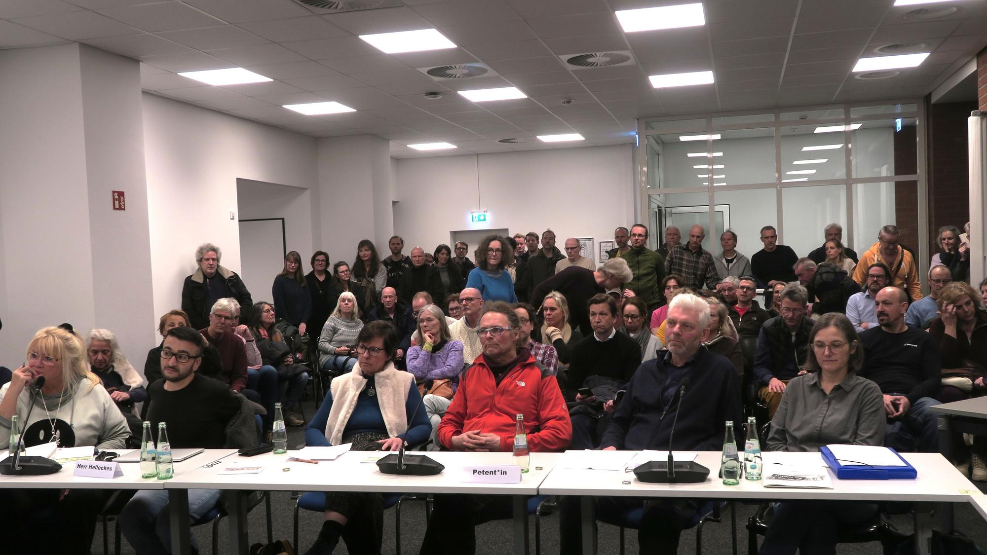An einem Tisch sitzen sechs Menschen, dahinter ist Publikum auf Stühlen und stehend zu sehen.
