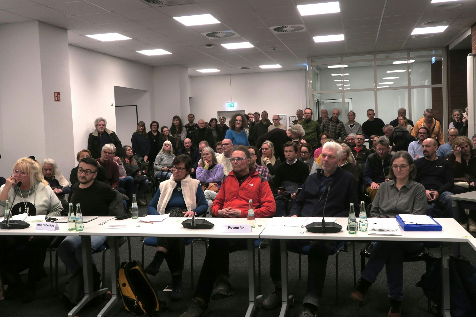 In einem Raum stehen viele Menschen, andere sitzen an Tischen oder in Zuschauerreihen.