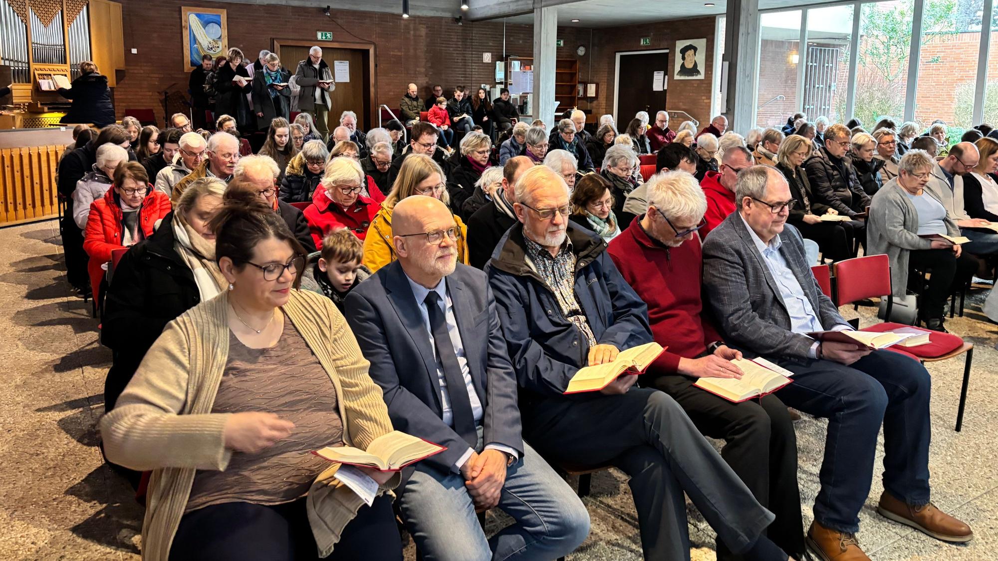In einer Kirche sitzen einige Gemeindemitgleider auf Stühlen und schauen auf Gesangsbücher.