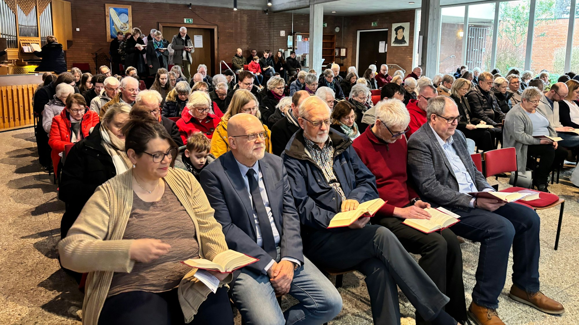 In einer Kirche sitzen einige Gemeindemitgleider auf Stühlen und schauen auf Gesangsbücher.