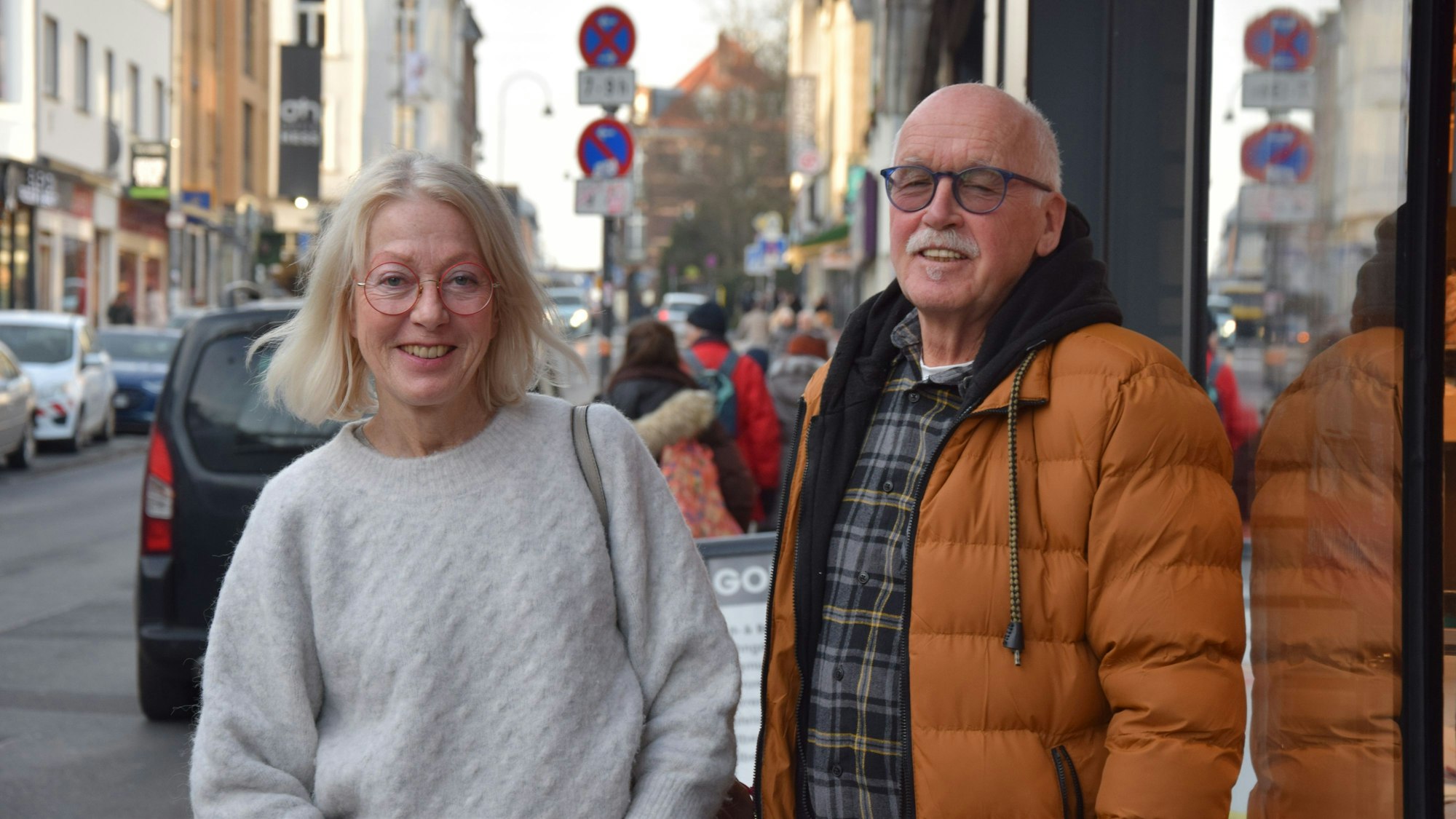 Eine Frau mit blondem Haar und ein Mann mit grauem Haar stehen auf einer belebten Straße.