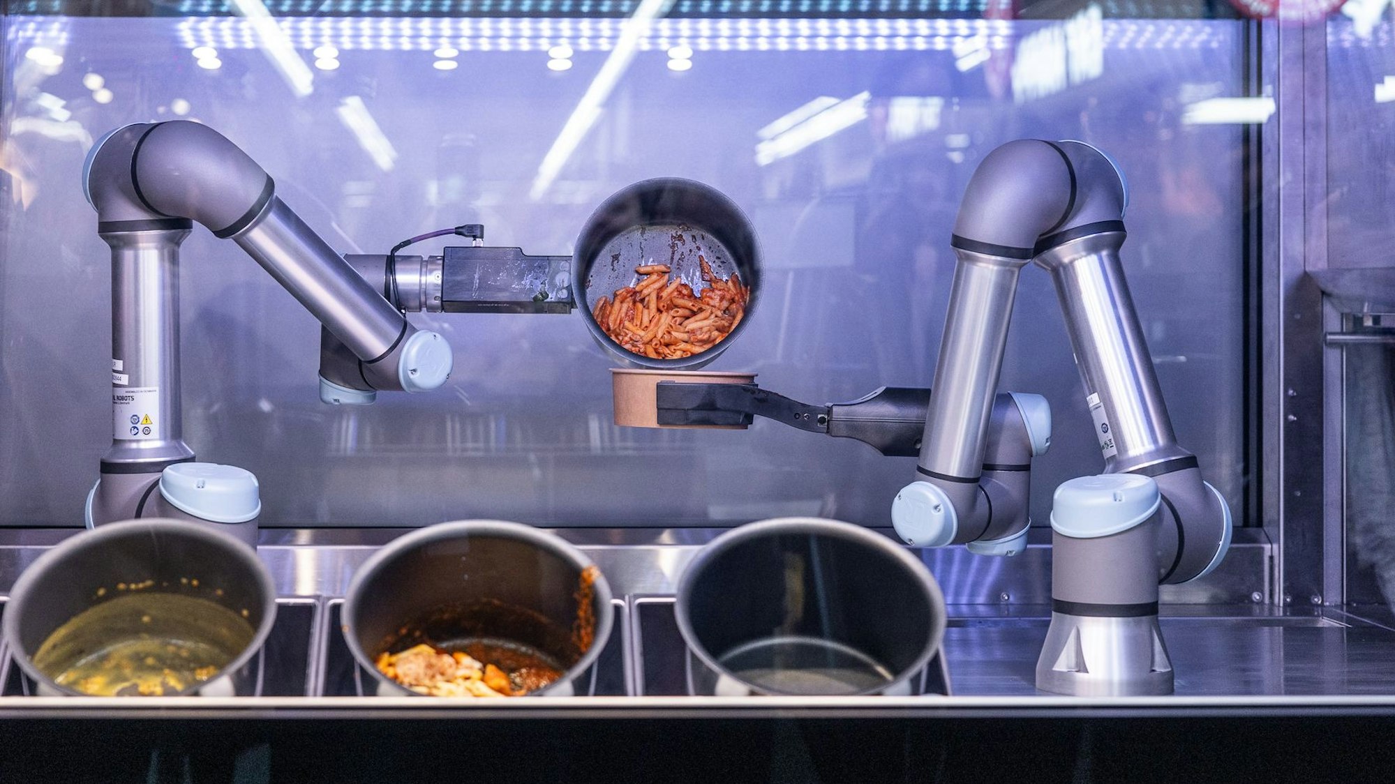 Zwei Roboterarme füllen heiße Nudeln aus einem Topf in eine Pappschale um.