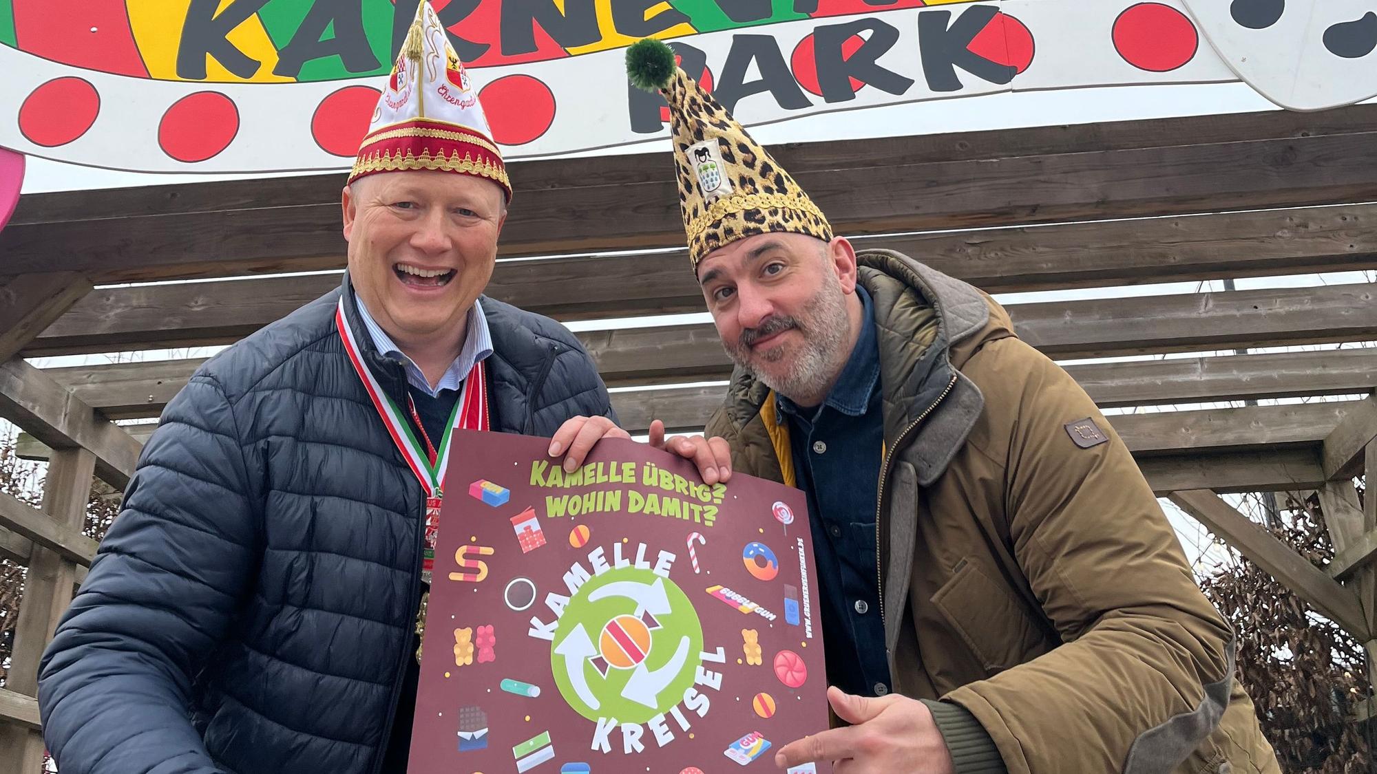 Das Foto zeigt den Landwirt und den Karnvalisten mit Karnevalsmützen und Plakat vor dem Karnevalspark auf dem Gertrudenhof.
