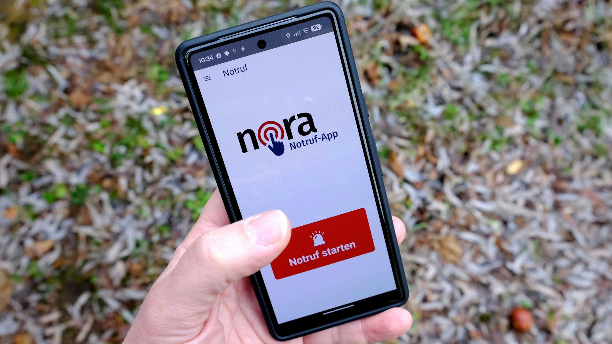 Auf dem Bildschirm eines Smartphones ist die Seite der App Nora zu sehen. Am unteren Rand hat sie ein rotes Feld, auf das man tippt, um einen Notruf zu senden.
