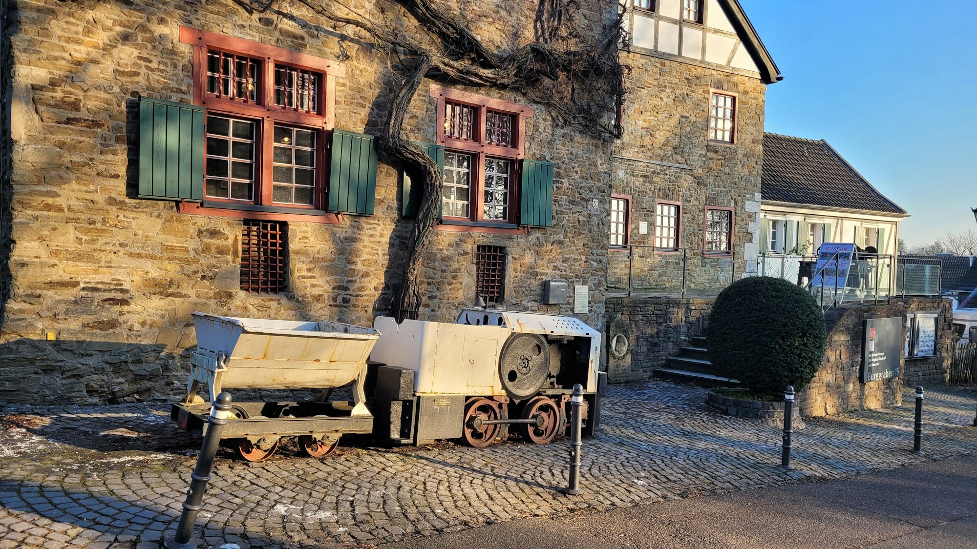 Ein altes Haus mit Bruchsteinmauerwerk und grünen Schlagläden. Davor eine Kohlenlore.