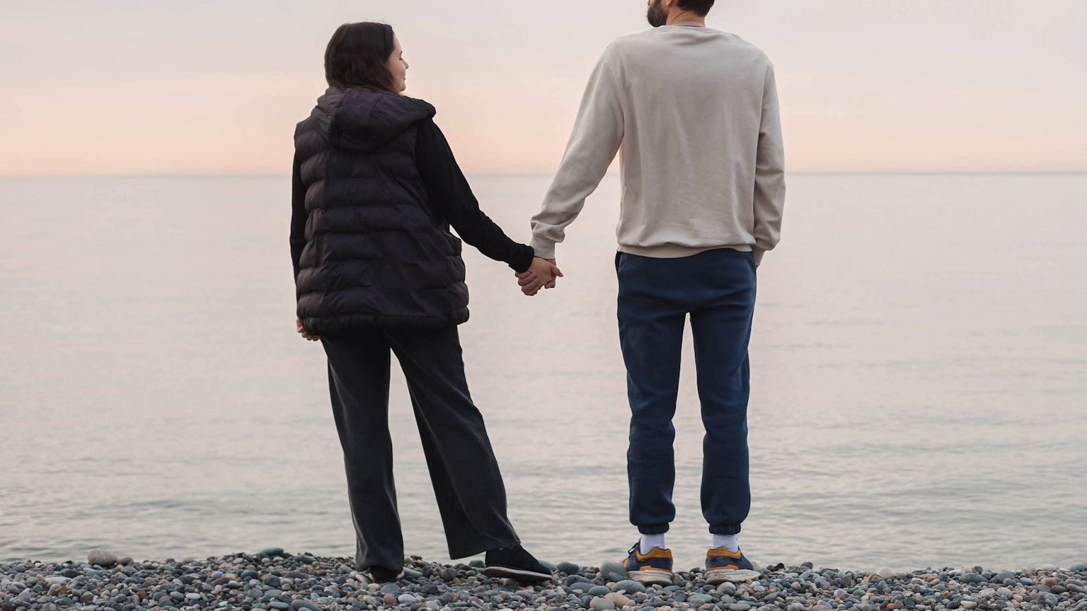 Paar stehen auf einem Kiesstrand, halten Hände und gegenüber dem ruhigen Meer während des weichen Sonnenuntergangs.