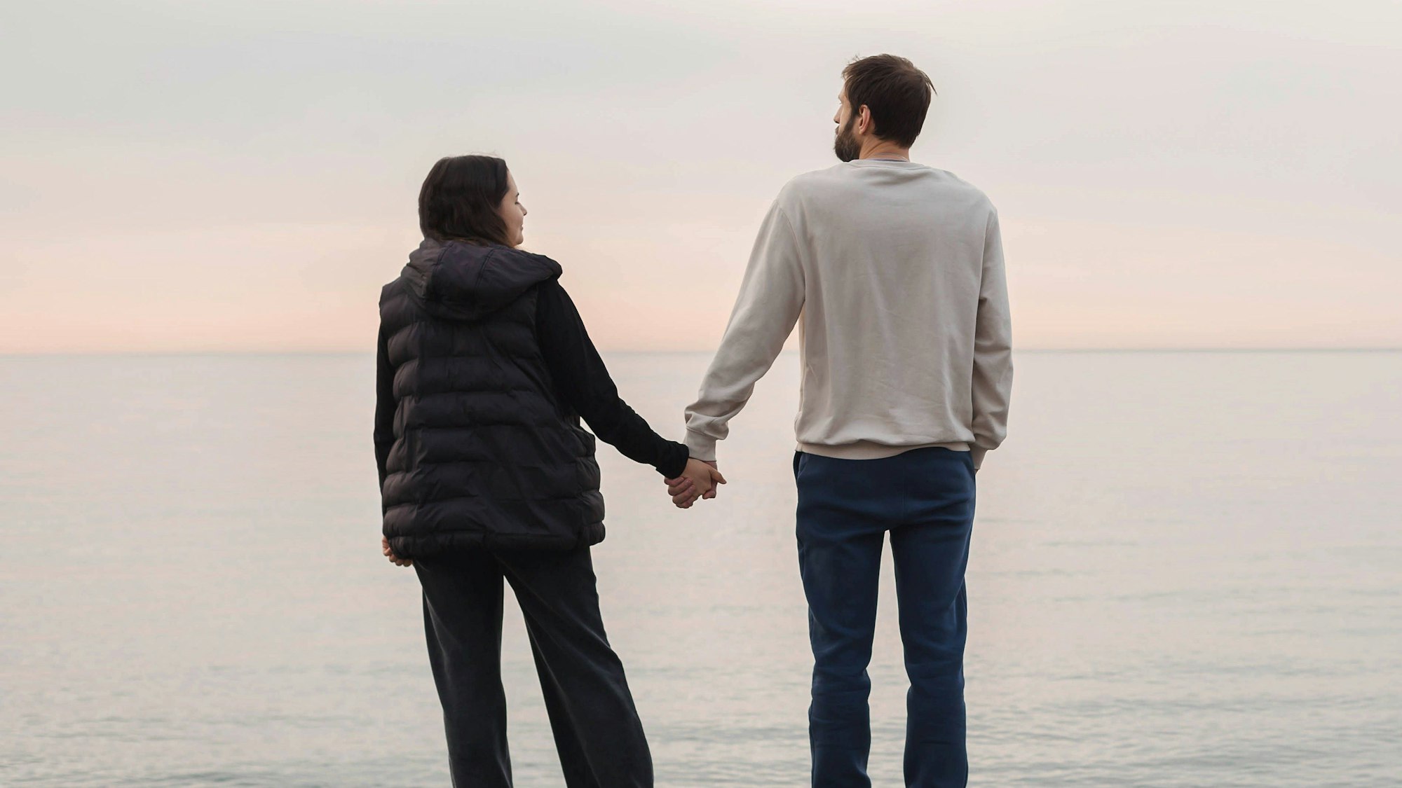 Paar stehen auf einem Kiesstrand, halten Hände und gegenüber dem ruhigen Meer während des weichen Sonnenuntergangs.