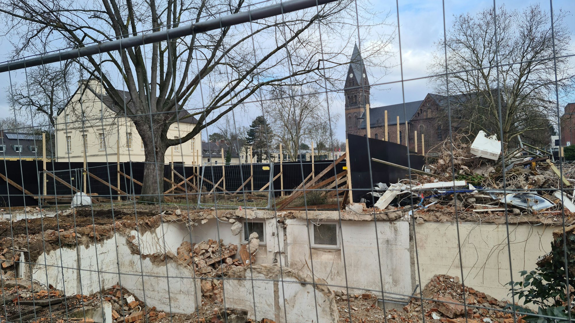 Hinter einem Bauzaun sind Reste von Gebäuden zu sehen, dahinter ein altes Gebäude und eine Kirche.