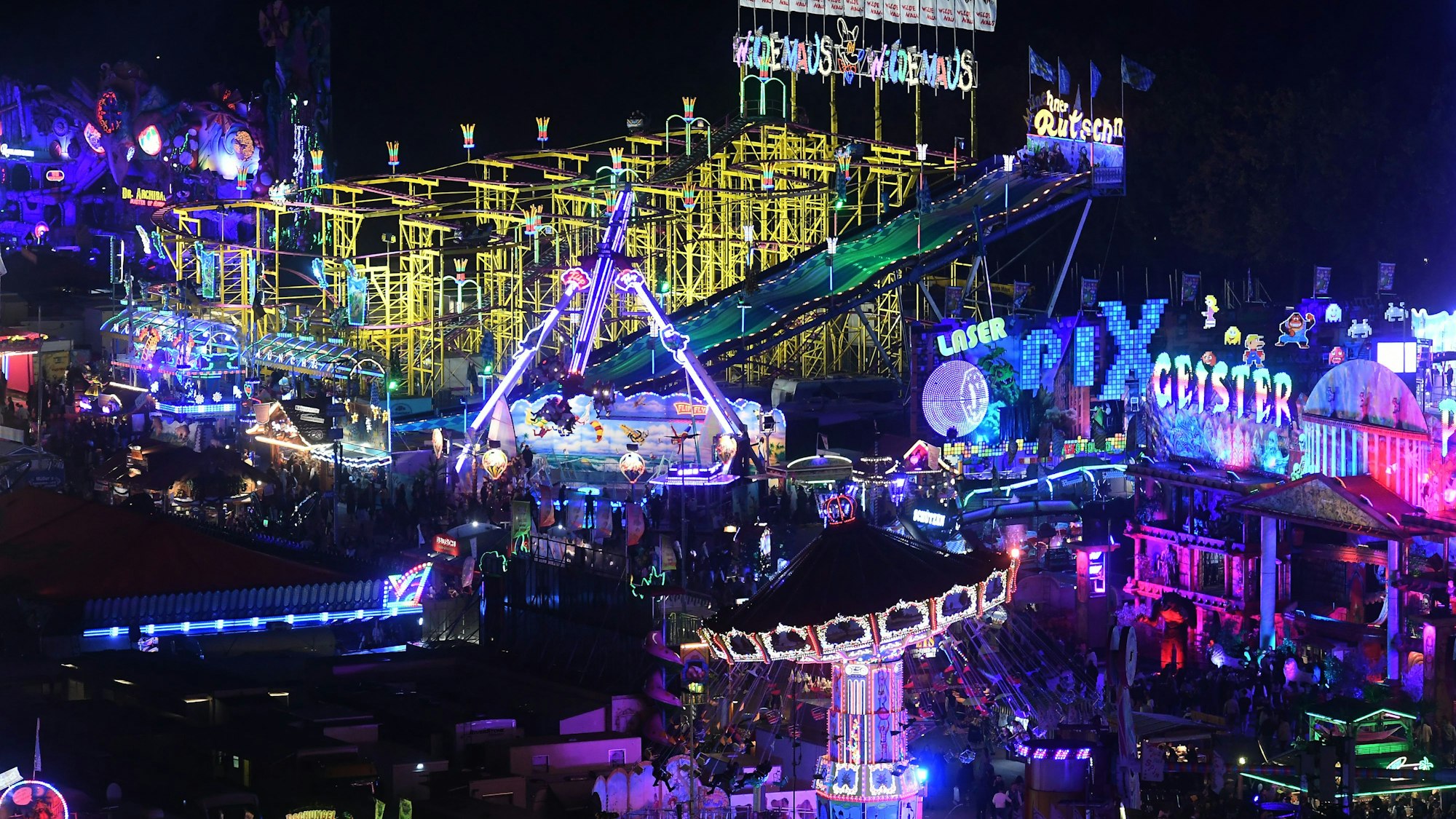 Beleuchtete Fahrgeschäfte auf dem Oktoberfest sind bei Dunkelheit aus dem Riesenrad zu sehen.