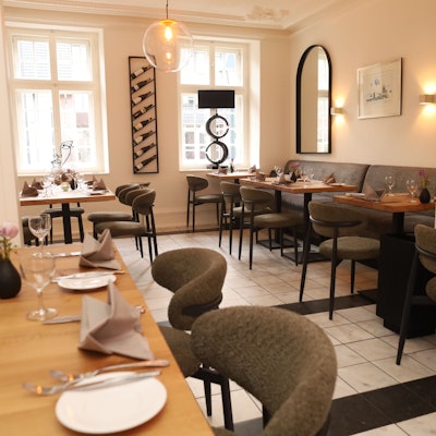 Der Gastraum ist zu sehen, er ist in weiß gehalten, Holztische und grau bezogene Sitzmöbel stehen in warmem Licht.