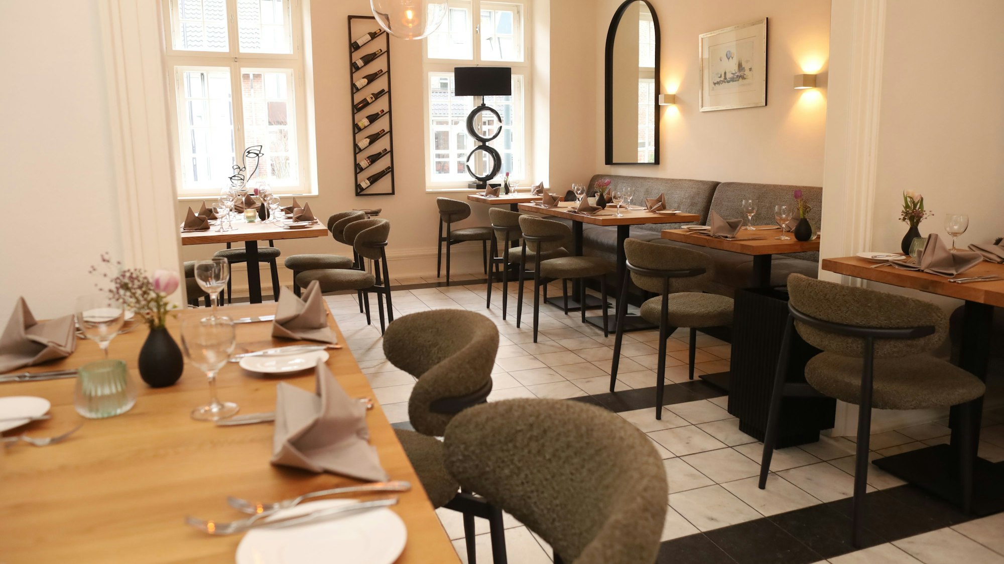 Der Gastraum ist zu sehen, er ist in weiß gehalten, Holztische und grau bezogene Sitzmöbel stehen in warmem Licht.