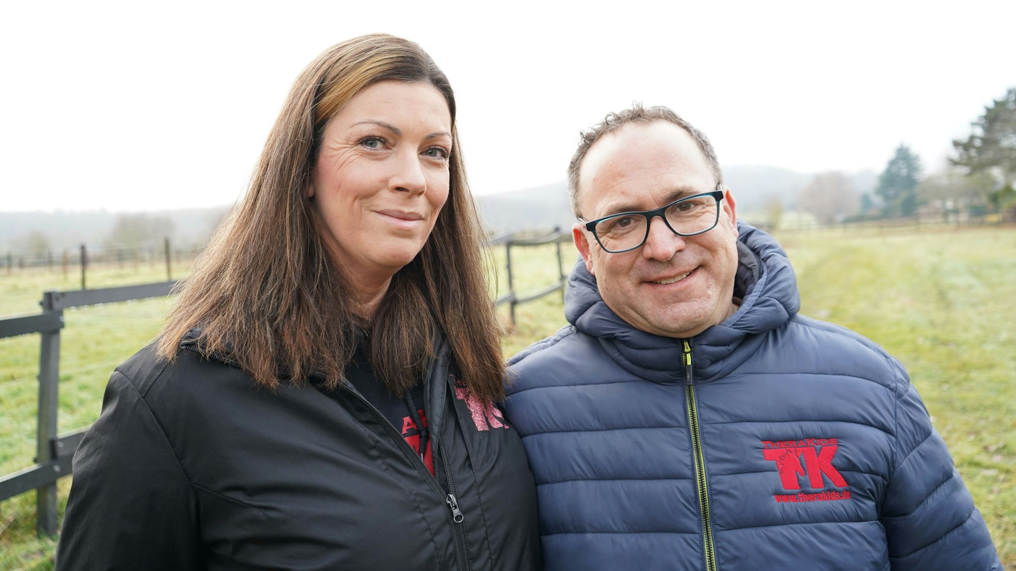 Die beiden tragen Jacken mit dem Vereinslogo, einem TK und der Aufschrift TheraKids. Im Hintergrund Wiesen und Zäune.