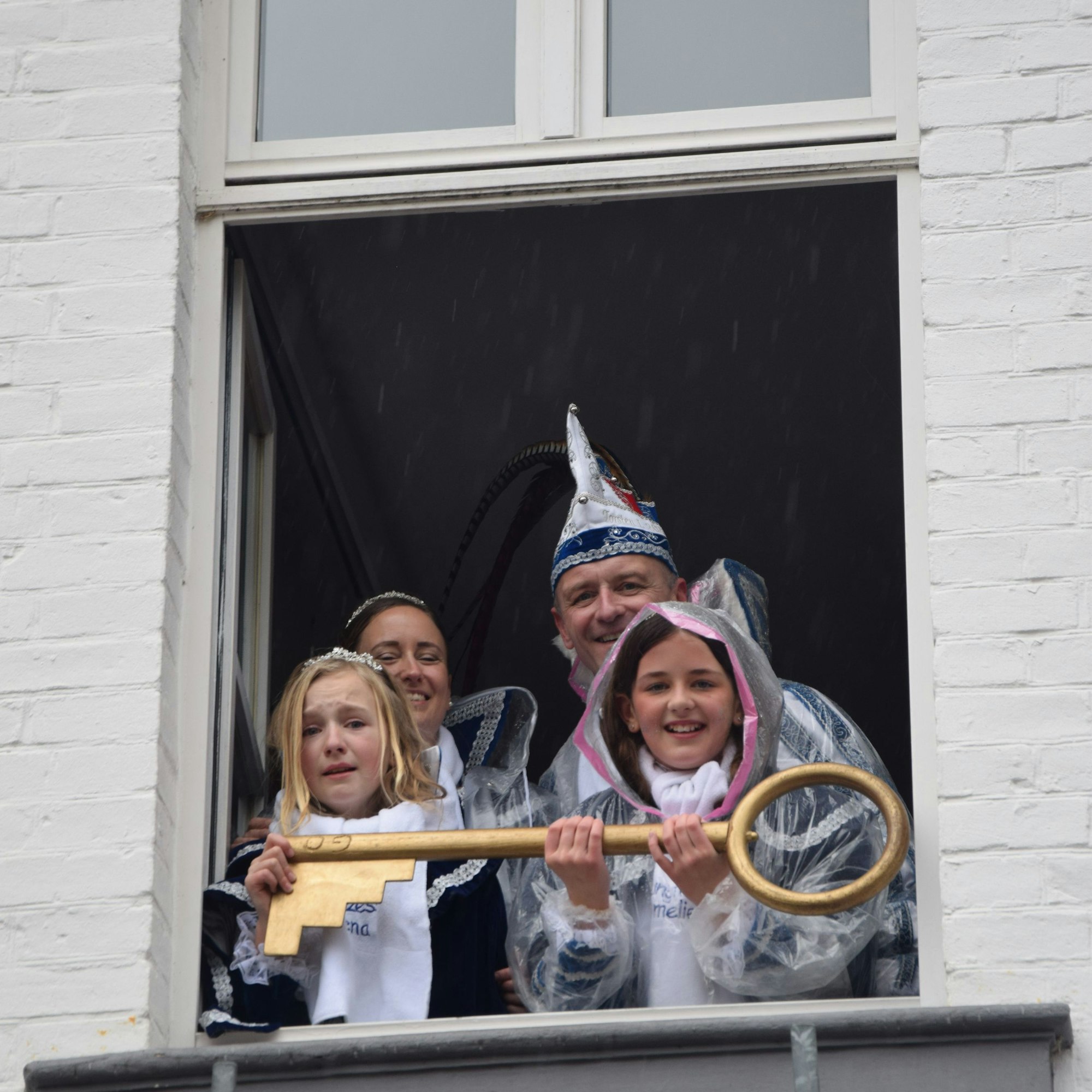 Zwei Kinderprinzessinnen und ein Prinzenpaar in blau-weißem Ornat schauen aus einem Fenster. In der Hand haben sie einen großen goldenen Schlüssel.