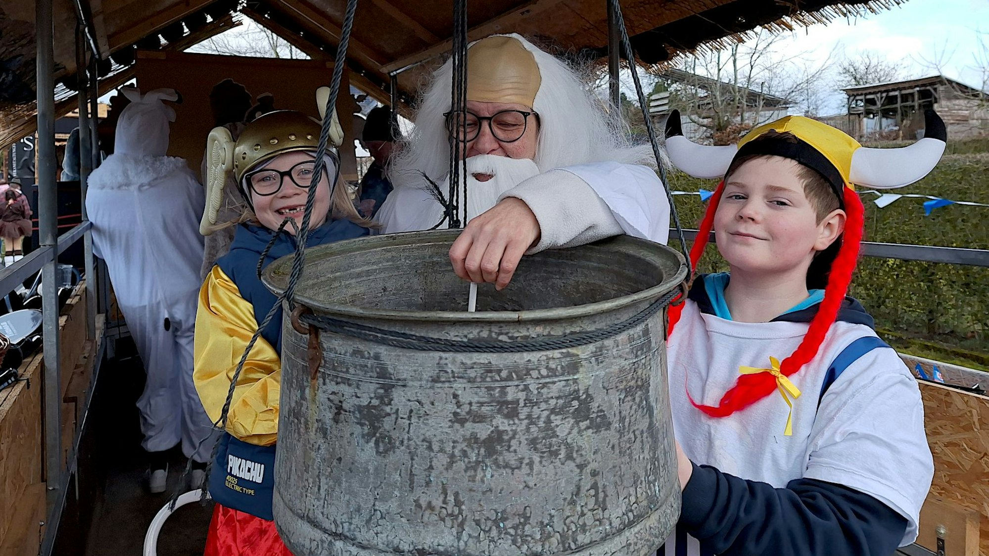 Eine als Miraculix verkleidete Frau rührt in einem Bottich. Links und rechts stehen Kinder, die als Asterix und Obelix verkleidet sind.