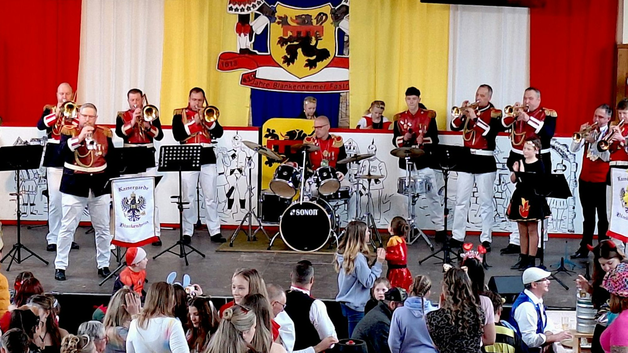 Die Musiker der Blankenheimer Kaisergarde stehen in rot-weißen Uniformen mit ihren Instrumenten auf der Bühne. Im Vordergrund ist Publikum zu sehen