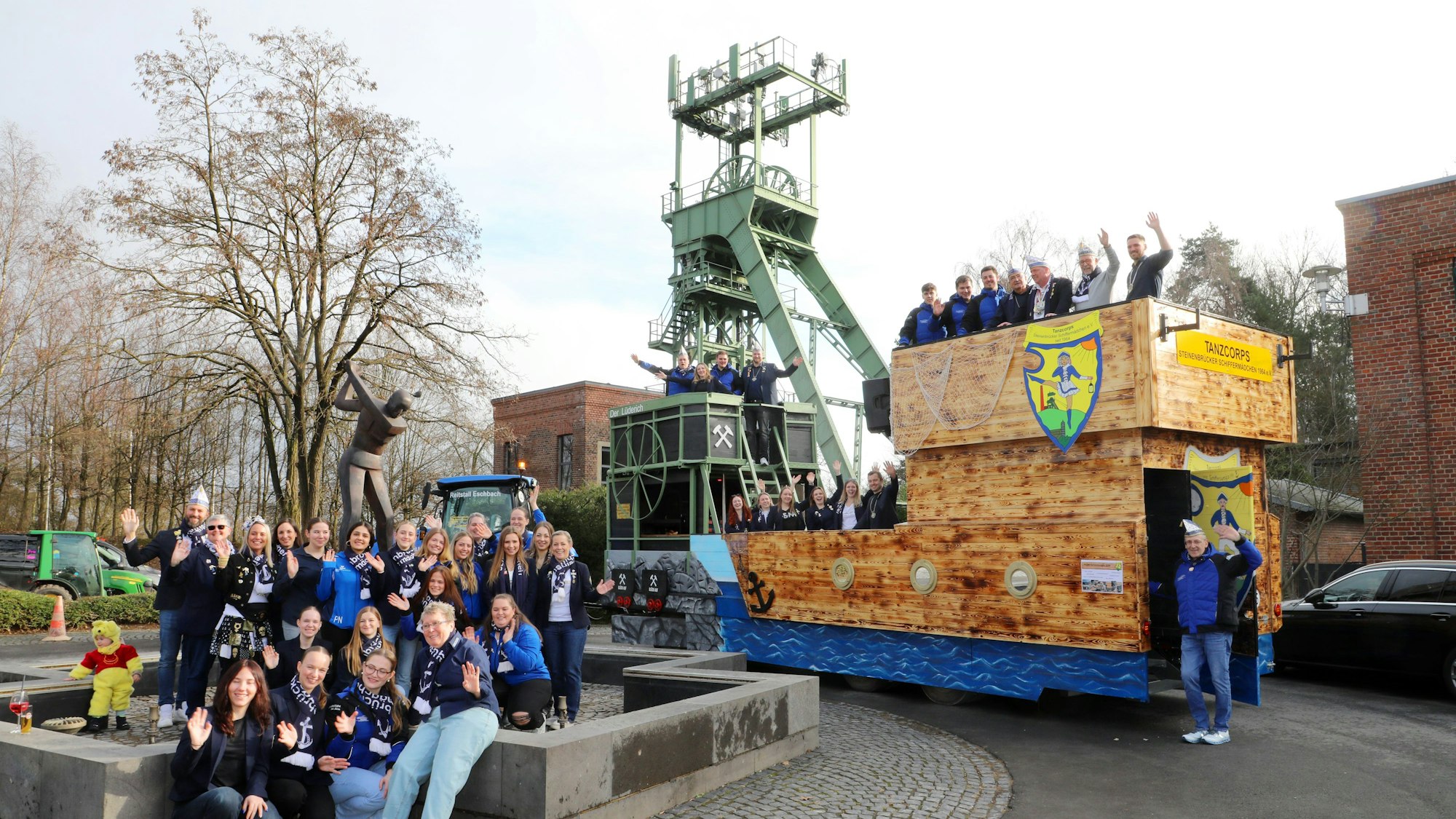 Links eine Gruppe von Menschen, rechts ein Karnevalswagen, der auf der linken Seite dem grünen Bergbau-Förderturm nachempfunden ist. Das Original ragt dahinter auf und bildet visuell ein Einheit mit dem Element vom Karnevalswagen, der für die Karnevalisten ein Podest aus hellem Holz hat.