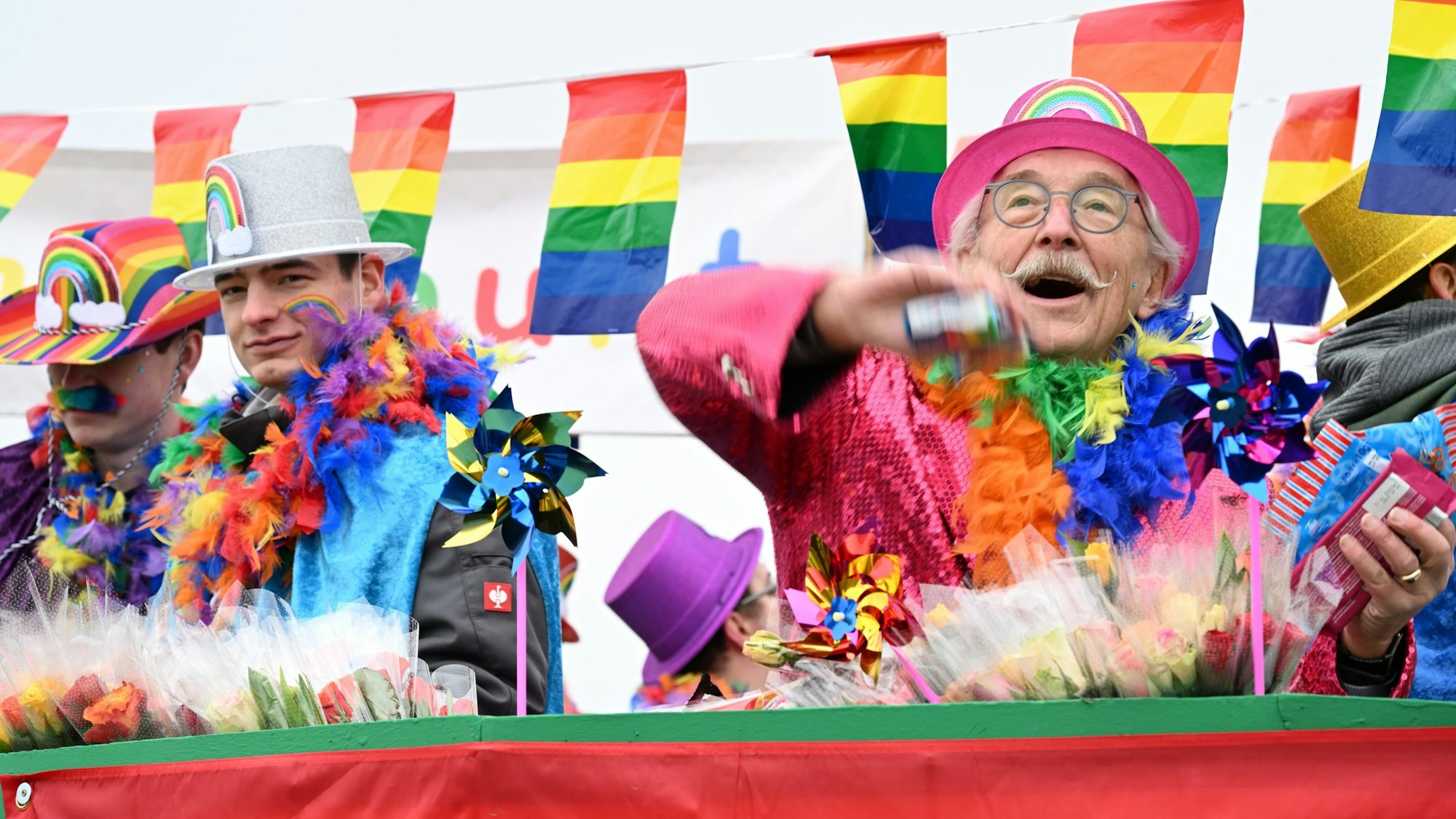 Karnevalisten, kostümiert in den Farben des Regenbogens, werfen Kamelle von einem Wagen.