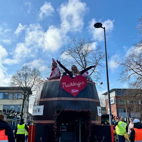 Aus einem Karnevalswagen, der wie ein riesiges Fass gebaut ist, schaut oben die Maikönigin heraus.
