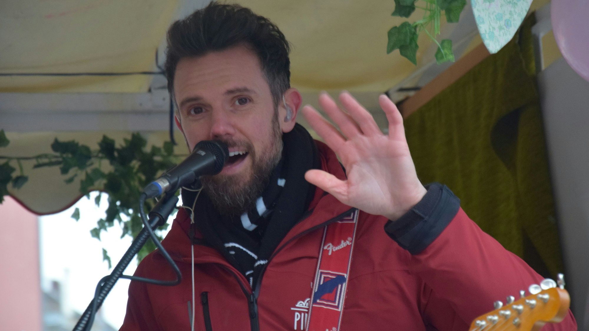 Ein Mann mit Bart steht winkend an einem Mikrofon.