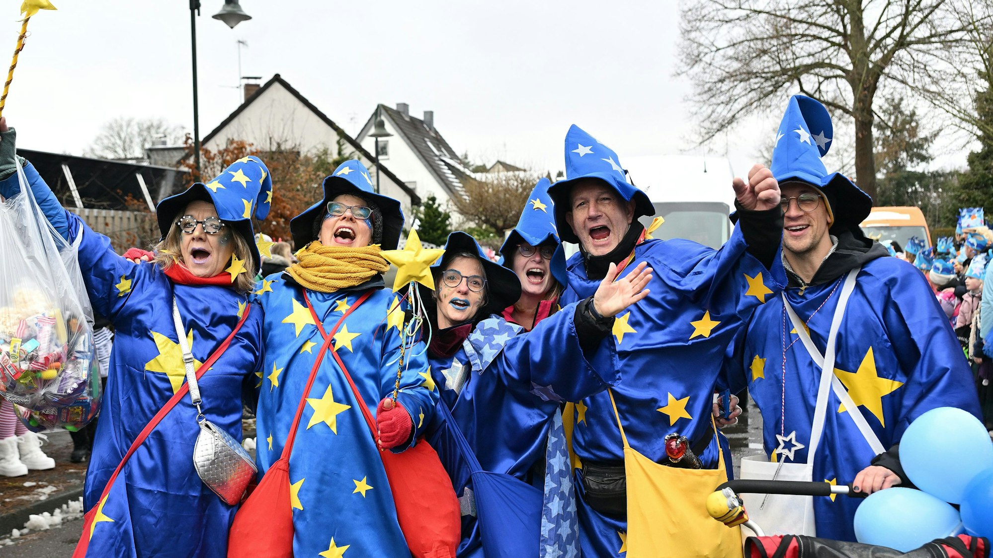 Sechs Erwachsene in himmelblauen Zauberermänteln und ebensolchen Hüten reißen die Arme hoch und rufen etwas.