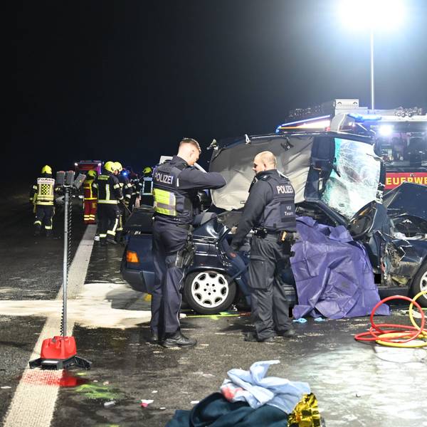 Zu sehen ist das Unfallwrack eines Autos, Polizisten stehen am Wagen, im Hintergrund steht ein Feuerwehrfahrzeug.