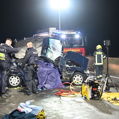 Zu sehen ist das Unfallwrack eines Autos, Polizisten stehen am Wagen, im Hintergrund steht ein Feuerwehrfahrzeug.
