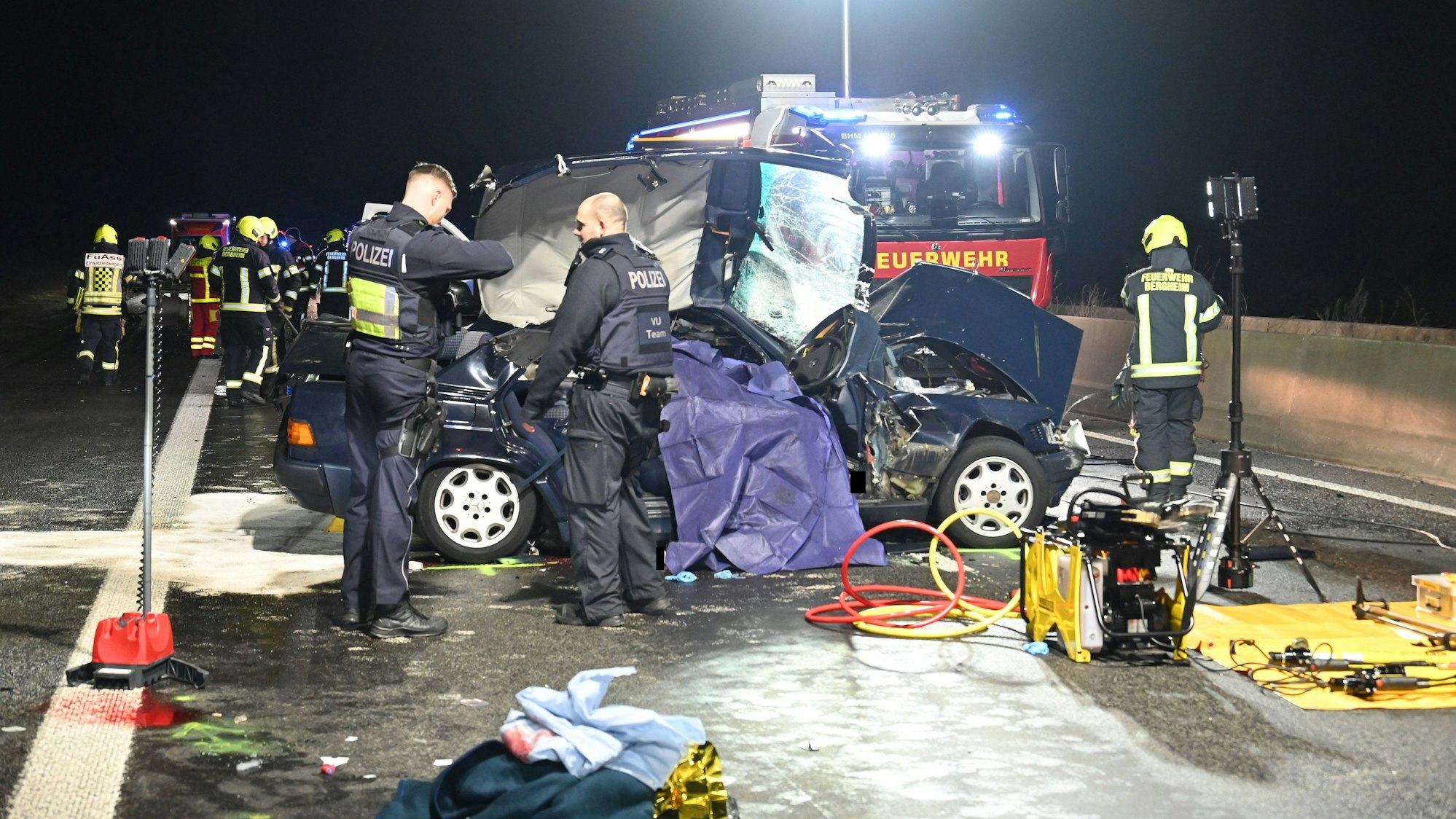 Zu sehen ist das Unfallwrack eines Autos, Polizisten stehen am Wagen, im Hintergrund steht ein Feuerwehrfahrzeug.