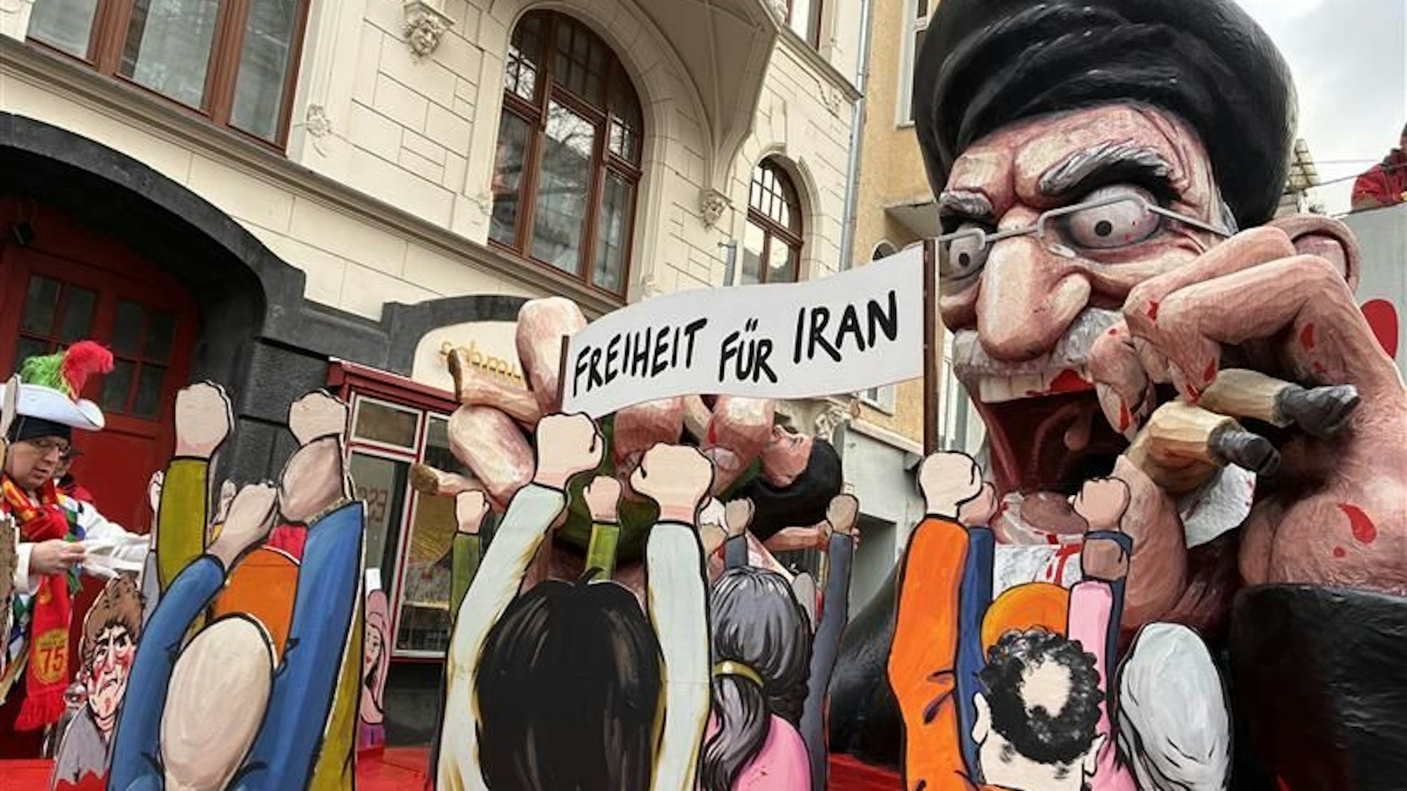 Das Bild zeigt den Wagen beim Kölner Rosenmontagszug, der den iranischen Politiker und Religionsführer Ali Chamenei zeigt. Blutrünstig verschlingt er sein Volk.