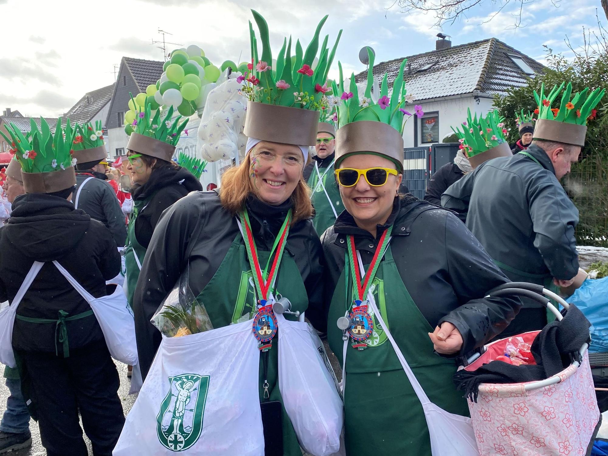 Zwei Frauen mit grünen Schürzen. Auf dem Kopf - aus Pappe - grünes Gras und bunte Blumen.