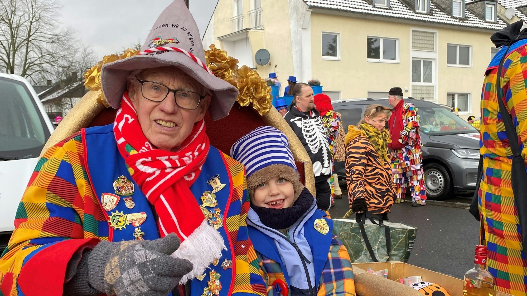 Ein alter Mann in buntem Mantel und rotem Schal neben einem kleinen Jungen. Beide schauen neugierig in die Kamera.