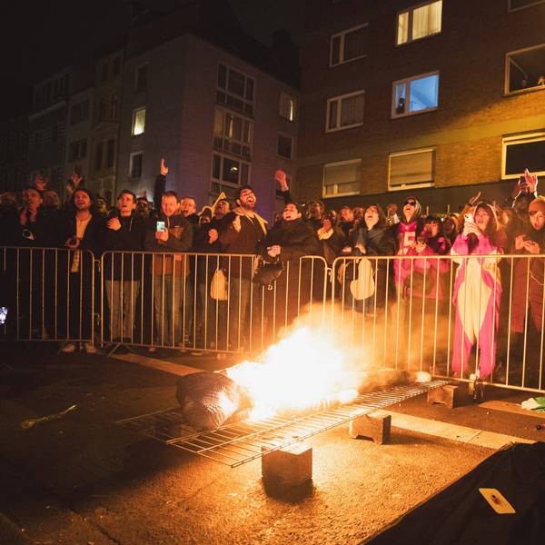 Ein brennender Nubbel liegt bei Nubbelverbrennung in der Schaafenstraße in Köln auf einem Gitter, dahinter feiern Karnevalisten hinter einer Absperrung. (Archivbild)