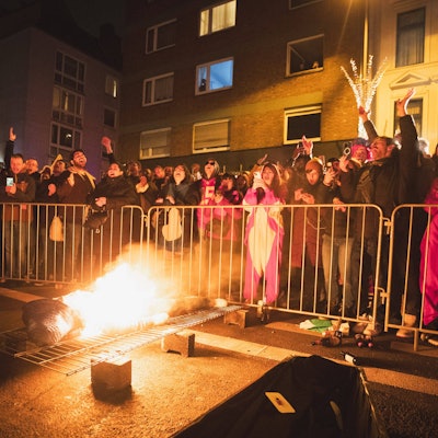 Ein brennender Nubbel liegt bei Nubbelverbrennung in der Schaafenstraße in Köln auf einem Gitter, dahinter feiern Karnevalisten hinter einer Absperrung. (Archivbild)