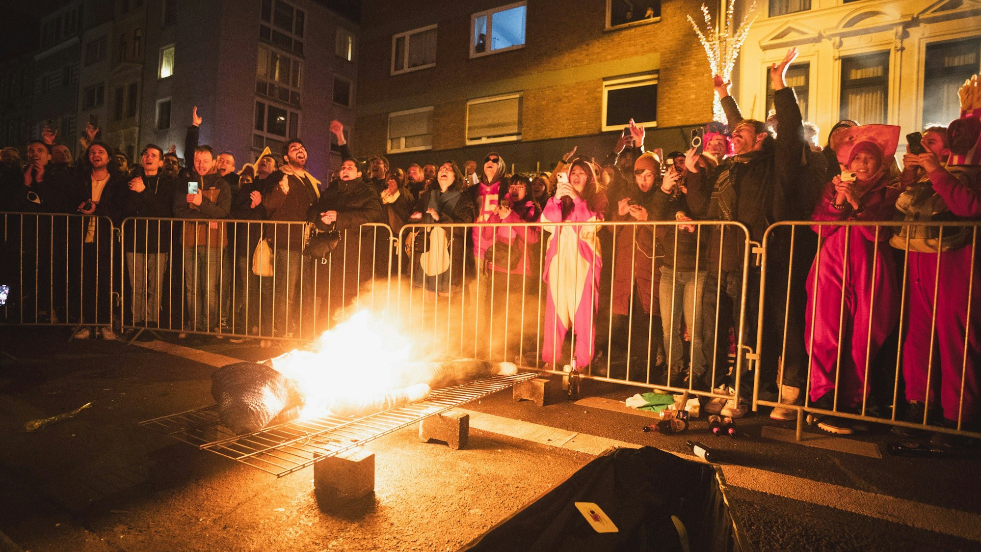 Ein brennender Nubbel liegt bei Nubbelverbrennung in der Schaafenstraße in Köln auf einem Gitter, dahinter feiern Karnevalisten hinter einer Absperrung. (Archivbild)