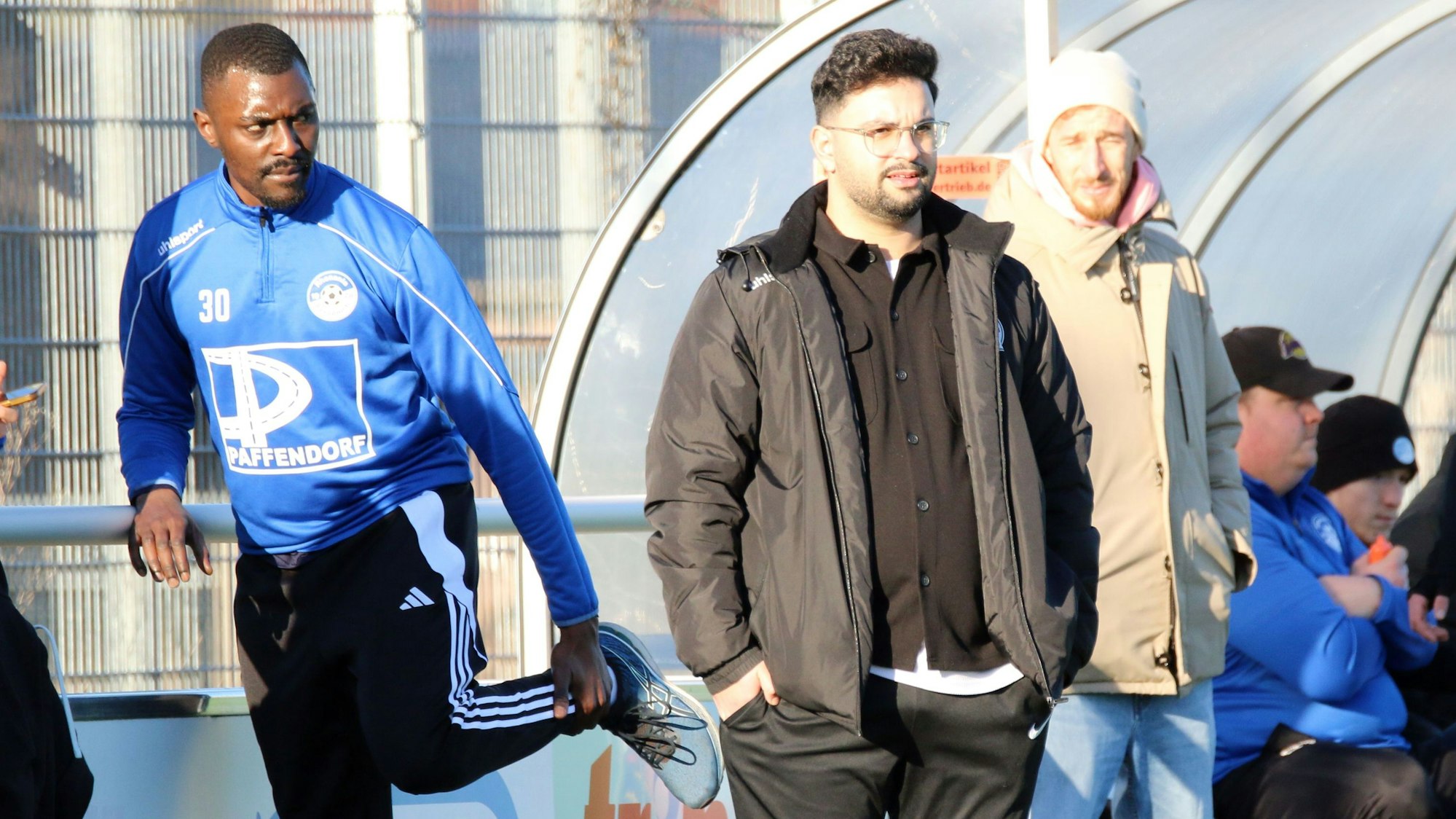 Bessenichs Trainer Can Celik verfolgt ein Spiel und hat die Hände in den Hosentaschen. Neben ihm macht sich ein Spieler warm. Dahinter steht Moritz Hartmann mit einer Mütze auf dem Kopf.