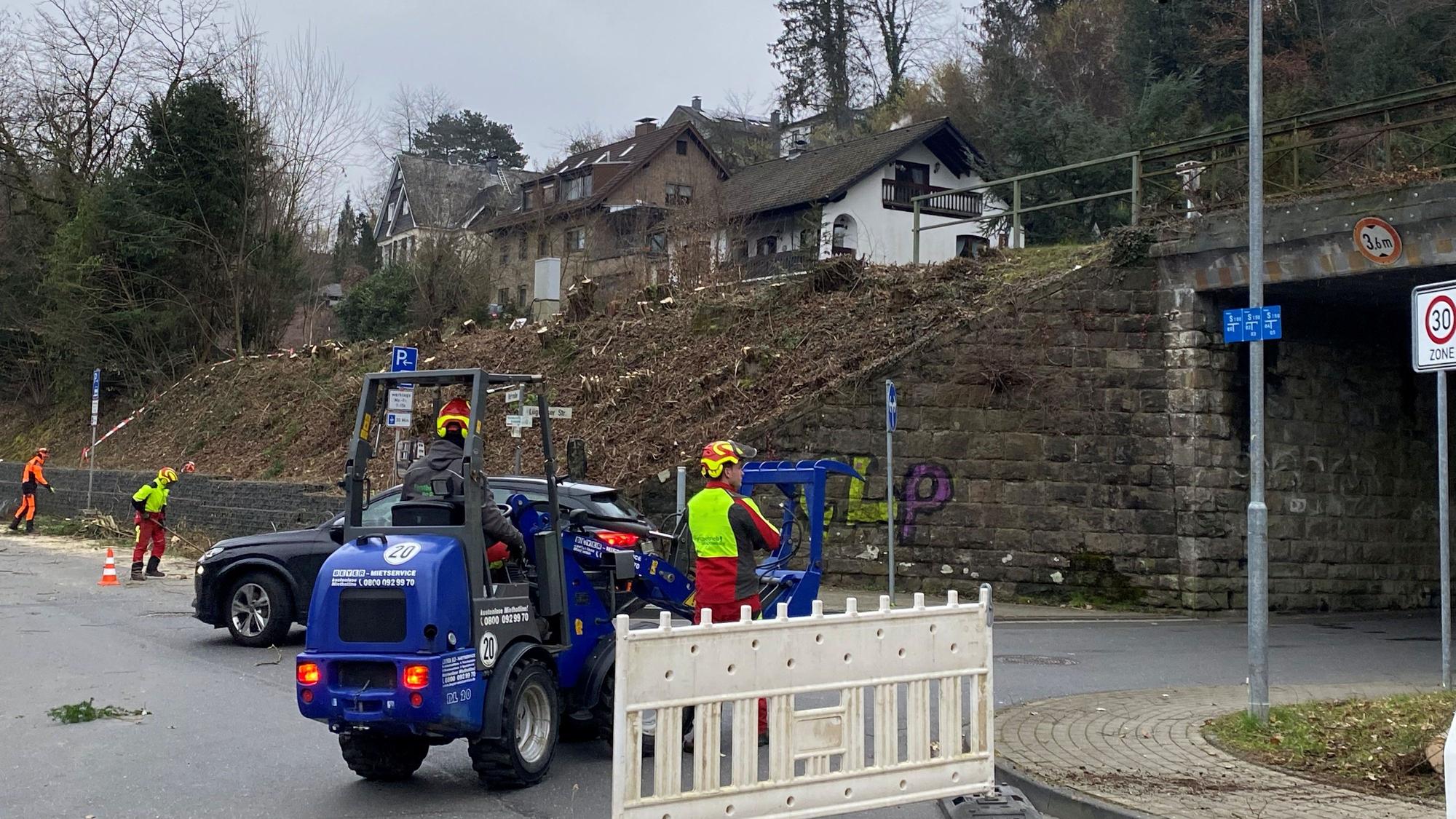 Eine Bahnunterführung, davor eine blaue Baumaschine und drei Arbeiter in roten Overalls. Der Hang neben der Unterführung ist gerodet, dort stehen nur noch Baumstümpfe.
