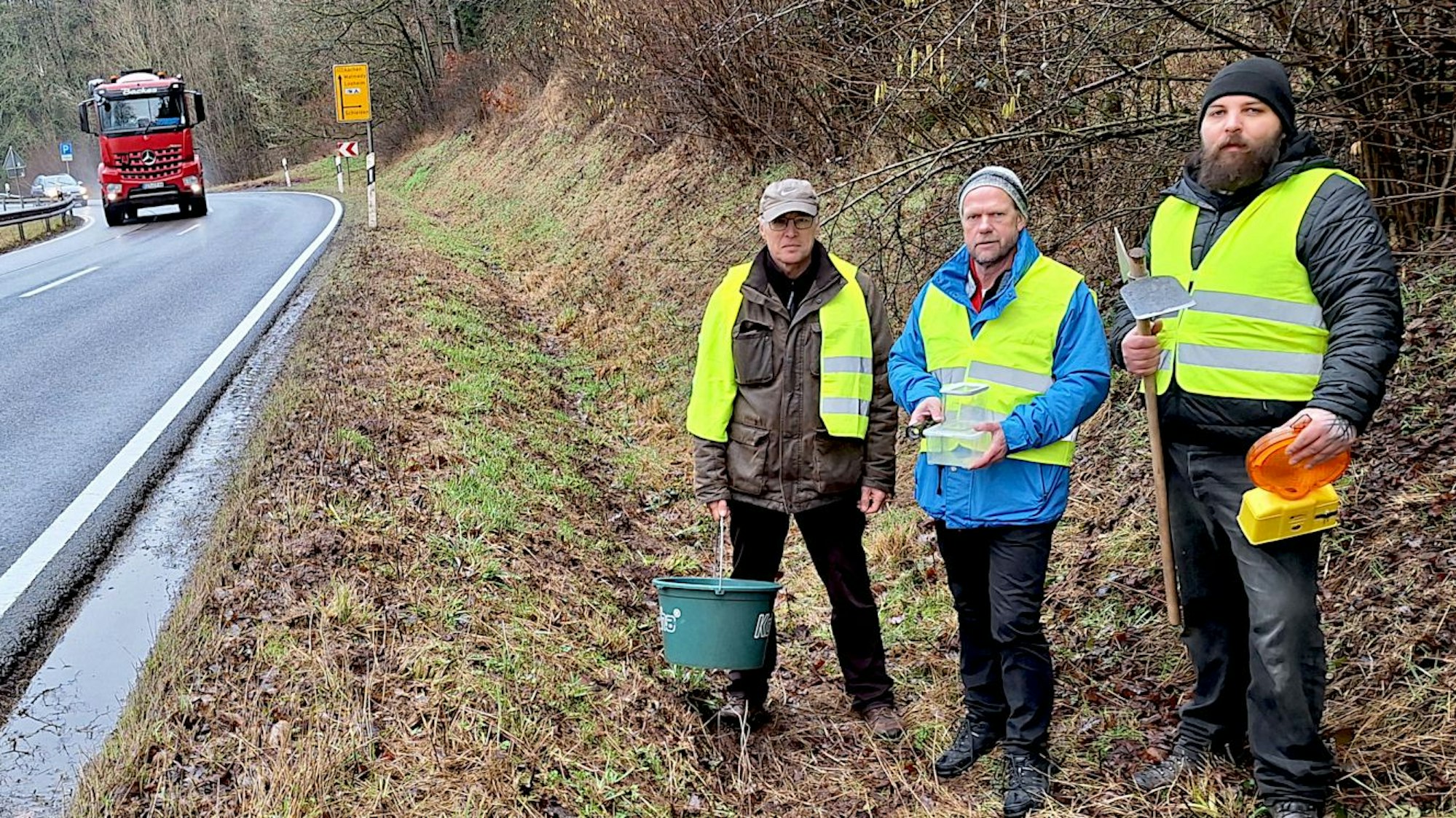 Drei Männer tragen gelbe Warnwesten und stehen an einer Landstraße, auf der gerade ein Lastwagen herangefahren kommt.