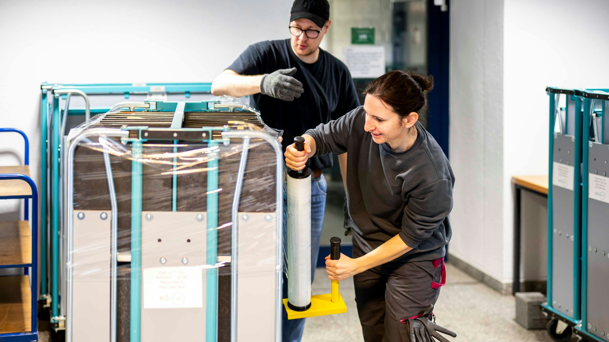 Das Bild zeigt Judith Hages, wie sie mit einer überdimensionalen Frischhaltefolie um den Rollcontainer geht.