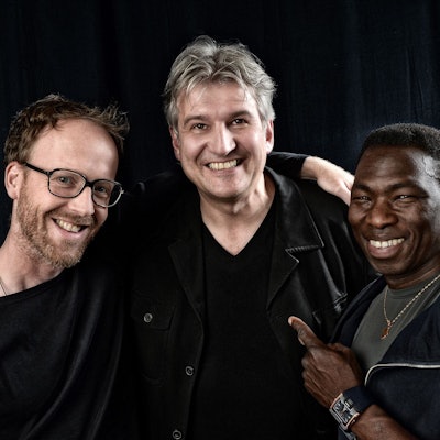 Drei Männer stehen Arm in Arm nebeneinander und lächeln in die Kamera.